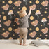 Lovely child standing in front of Whimsy Tots Moonlit Bear Dreams Wallpaper in Midnight Cocoa Brown with bear, flower, moon, star in design. Lovely child standing in front of Whimsy Tots Moonlit Bear Dreams Wallpaper in Midnight Cocoa Brown with bear, flower, moon, star in design.