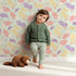Lovely child standing in front of Whimsy Tots Pastel Feather Garden Wallpaper in Mint Whisper Green with flower, leaf, feather, petal in design. Lovely child standing in front of Whimsy Tots Pastel Feather Garden Wallpaper in Mint Whisper Green with flower, leaf, feather, petal in design.
