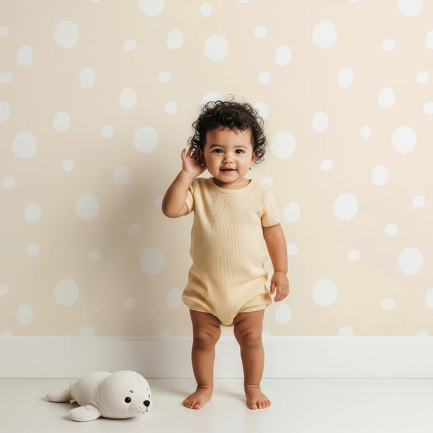 Lovely child standing in front of Whimsy Tots Gentle Pebble Dots Wallpaper in Butter Beige with dot, spot, pebble, beige in design.