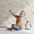 Lovely child standing in front of Whimsy Tots Wildflower Scribble Garden Wallpaper in Cream Sketch with flower, leaf, berry, sketch, doodle in design. Lovely child standing in front of Whimsy Tots Wildflower Scribble Garden Wallpaper in Cream Sketch with flower, leaf, berry, sketch, doodle in design.