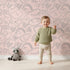 Lovely child standing in front of Whimsy Tots Doodle Meadow Shapes Wallpaper in Blush Pink with doodle, shape, scribble, petal, arch in design. Lovely child standing in front of Whimsy Tots Doodle Meadow Shapes Wallpaper in Blush Pink with doodle, shape, scribble, petal, arch in design.