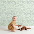 Lovely child standing in front of Whimsy Tots Doodle Meadow Shapes Wallpaper in Mint Green with doodle, shape, petal, scribble, arch in design. Lovely child standing in front of Whimsy Tots Doodle Meadow Shapes Wallpaper in Mint Green with doodle, shape, petal, scribble, arch in design.