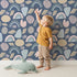 Lovely child standing in front of Whimsy Tots Rainbow Petal Shapes Wallpaper in Midnight Blue with rainbow, petal, shape, arch, stripe in design. Lovely child standing in front of Whimsy Tots Rainbow Petal Shapes Wallpaper in Midnight Blue with rainbow, petal, shape, arch, stripe in design.