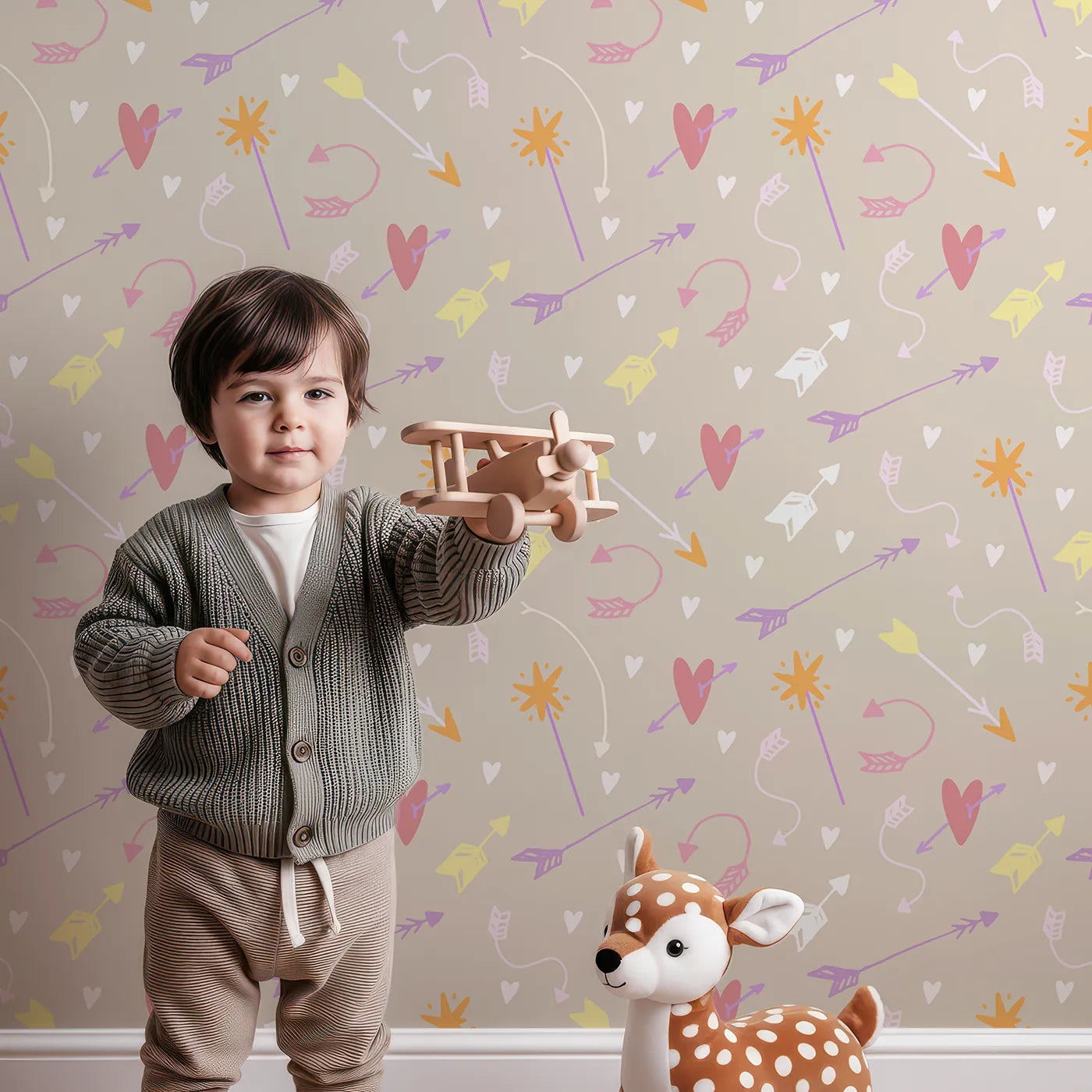 Lovely child standing in front of Whimsy Tots Cupids Arrow Parade Wallpaper in Cozy Beige with arrow, heart, star in design.