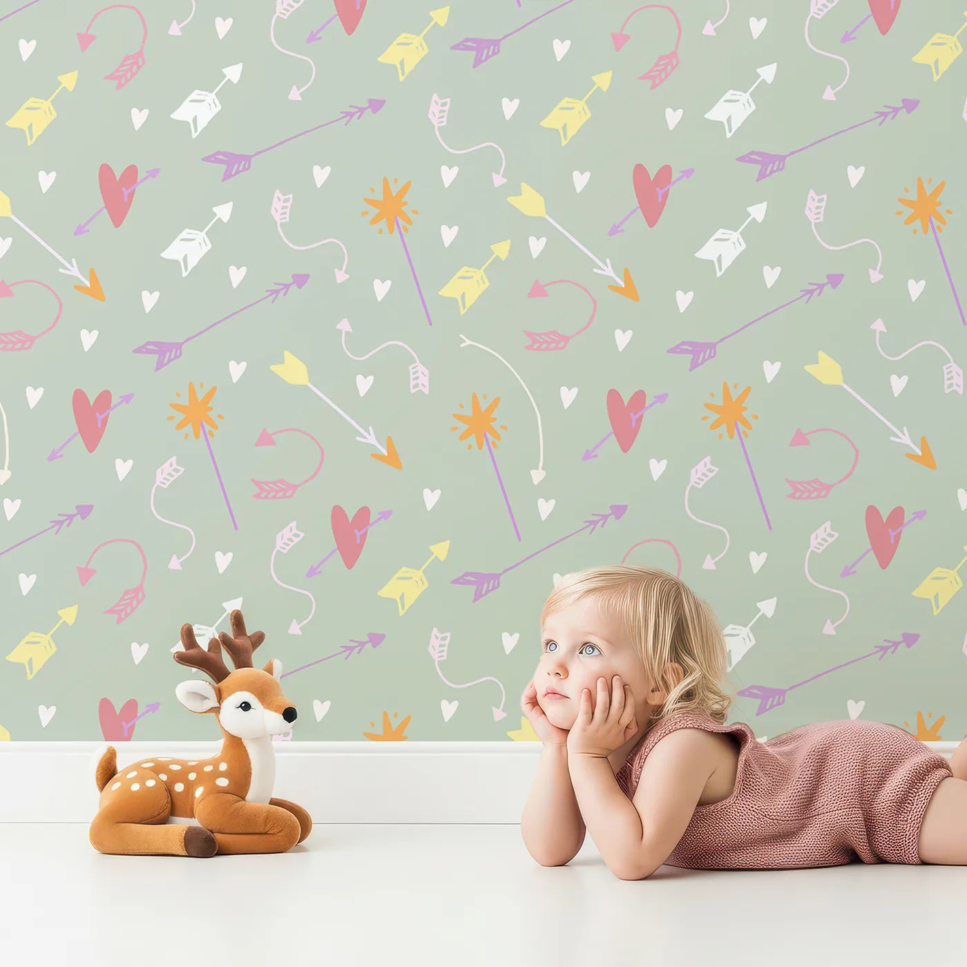 Lovely child standing in front of Whimsy Tots Cupids Arrow Parade Wallpaper in Sage Whimsy Green with arrow, heart, star in design.