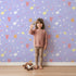 Lovely child standing in front of Whimsy Tots Cupids Arrow Parade Wallpaper in Lavender Pop with arrow, heart, star in design. Lovely child standing in front of Whimsy Tots Cupids Arrow Parade Wallpaper in Lavender Pop with arrow, heart, star in design.