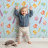 Lovely child standing in front of Whimsy Tots Feathered Joy Parade Wallpaper in Sky Blue with bird, feather, sprig, arrow in design. Lovely child standing in front of Whimsy Tots Feathered Joy Parade Wallpaper in Sky Blue with bird, feather, sprig, arrow in design.