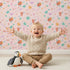 Lovely child standing in front of Whimsy Tots Love and Arrows Bloom Parade Wallpaper in Blush Pink with arrow, heart, flower, burst in design. Lovely child standing in front of Whimsy Tots Love and Arrows Bloom Parade Wallpaper in Blush Pink with arrow, heart, flower, burst in design.