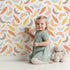 Lovely child standing in front of Whimsy Tots Feathered Whimsy Drift Wallpaper in Vanilla Cream with feather, diamond, dot in design. Lovely child standing in front of Whimsy Tots Feathered Whimsy Drift Wallpaper in Vanilla Cream with feather, diamond, dot in design.