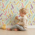 Lovely child standing in front of Whimsy Tots Feather Arrow Confetti Wallpaper in Light Mint Green with arrow, feather, triangle, dot in design. Lovely child standing in front of Whimsy Tots Feather Arrow Confetti Wallpaper in Light Mint Green with arrow, feather, triangle, dot in design.
