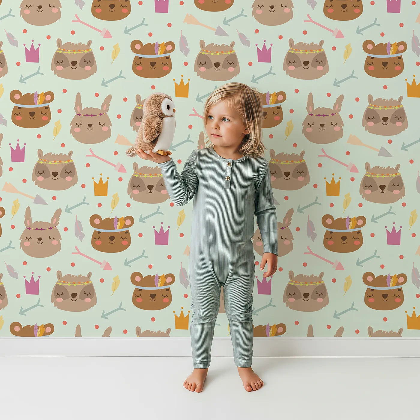 Lovely child standing in front of Whimsy Tots Feather Crown Critters Wallpaper in Meadow Green with bear, rabbit, crown, feather, arrow, dot in design.