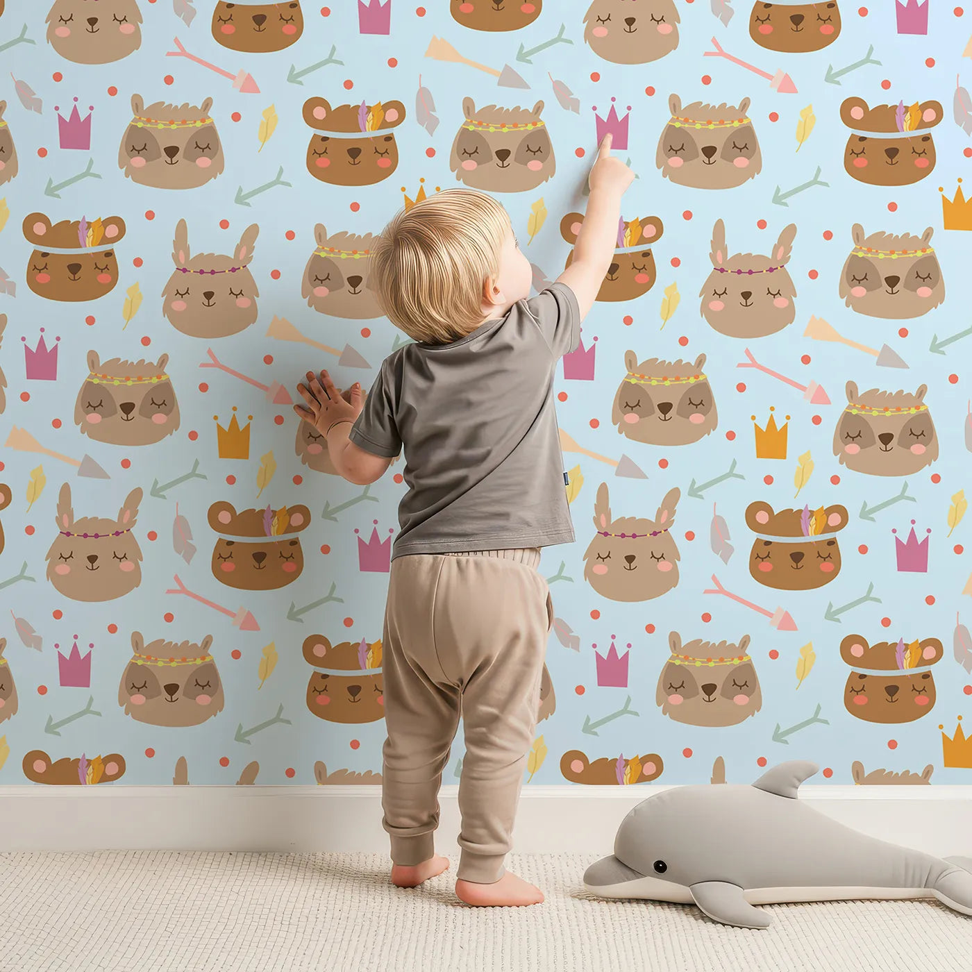 Lovely child standing in front of Whimsy Tots Feather Crown Critters Wallpaper in Soft Sky Blue with bear, rabbit, crown, feather, arrow, dot in design.