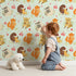 Lovely child standing in front of Whimsy Tots Cuddle Buddies Parade Wallpaper in Mint Green with fox, bird, badger, cat, heart, flower in design. Lovely child standing in front of Whimsy Tots Cuddle Buddies Parade Wallpaper in Mint Green with fox, bird, badger, cat, heart, flower in design.