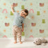 Lovely child standing in front of Whimsy Tots Garden Hop and Snail Trail Wallpaper in Mint Green with frog, snail, dragonfly, rainbow, sun, flower in design. Lovely child standing in front of Whimsy Tots Garden Hop and Snail Trail Wallpaper in Mint Green with frog, snail, dragonfly, rainbow, sun, flower in design.