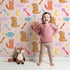 Lovely child standing in front of Whimsy Tots Arrow Pup and Foxy Night Parade Wallpaper in Blush Pink with dog, fox, moon, arrow, star, cloud in design. Lovely child standing in front of Whimsy Tots Arrow Pup and Foxy Night Parade Wallpaper in Blush Pink with dog, fox, moon, arrow, star, cloud in design.