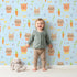 Lovely child standing in front of Whimsy Tots Meadow Crown Friends Wallpaper in Blue with fox, rabbit, bear, flower, carrot, crown, bow in design. Lovely child standing in front of Whimsy Tots Meadow Crown Friends Wallpaper in Blue with fox, rabbit, bear, flower, carrot, crown, bow in design.