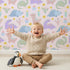 Lovely child standing in front of Whimsy Tots Cactus Dino Parade Wallpaper in Blush Pink with dinosaur, cactus, flower in design. Lovely child standing in front of Whimsy Tots Cactus Dino Parade Wallpaper in Blush Pink with dinosaur, cactus, flower in design.