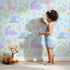 Lovely child standing in front of Whimsy Tots Cactus Dino Parade Wallpaper in Blue with dinosaur, cactus, flower in design. Lovely child standing in front of Whimsy Tots Cactus Dino Parade Wallpaper in Blue with dinosaur, cactus, flower in design.
