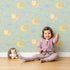 Lovely child standing in front of Whimsy Tots Snail Meadow Cuddle Parade Wallpaper in Sage Green with snail, flower, leaf in design. Lovely child standing in front of Whimsy Tots Snail Meadow Cuddle Parade Wallpaper in Sage Green with snail, flower, leaf in design.
