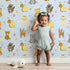 Lovely child standing in front of Whimsy Tots Magic Meadow Friends Wallpaper in Sky Blue with deer, koala, leopard, lion, raccoon, snail, arrow, wand, heart in design. Lovely child standing in front of Whimsy Tots Magic Meadow Friends Wallpaper in Sky Blue with deer, koala, leopard, lion, raccoon, snail, arrow, wand, heart in design.