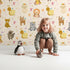 Lovely child standing in front of Whimsy Tots Magic Meadow Friends Wallpaper in Cream with deer, koala, leopard, lion, raccoon, snail, arrow, wand, heart in design. Lovely child standing in front of Whimsy Tots Magic Meadow Friends Wallpaper in Cream with deer, koala, leopard, lion, raccoon, snail, arrow, wand, heart in design.