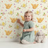 Lovely child standing in front of Whimsy Tots Tweeting Lovebirds Wallpaper in Cream with bird, owl, flower, heart, branch in design. Lovely child standing in front of Whimsy Tots Tweeting Lovebirds Wallpaper in Cream with bird, owl, flower, heart, branch in design.