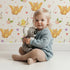 Lovely child standing in front of Whimsy Tots Tweeting Lovebirds Wallpaper in Soft White with bird, owl, flower, heart, branch in design. Lovely child standing in front of Whimsy Tots Tweeting Lovebirds Wallpaper in Soft White with bird, owl, flower, heart, branch in design.