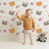 Lovely child standing in front of Whimsy Tots Meadow Snuggle Faces Wallpaper in White with cat, raccoon, flower, heart in design. Lovely child standing in front of Whimsy Tots Meadow Snuggle Faces Wallpaper in White with cat, raccoon, flower, heart in design.