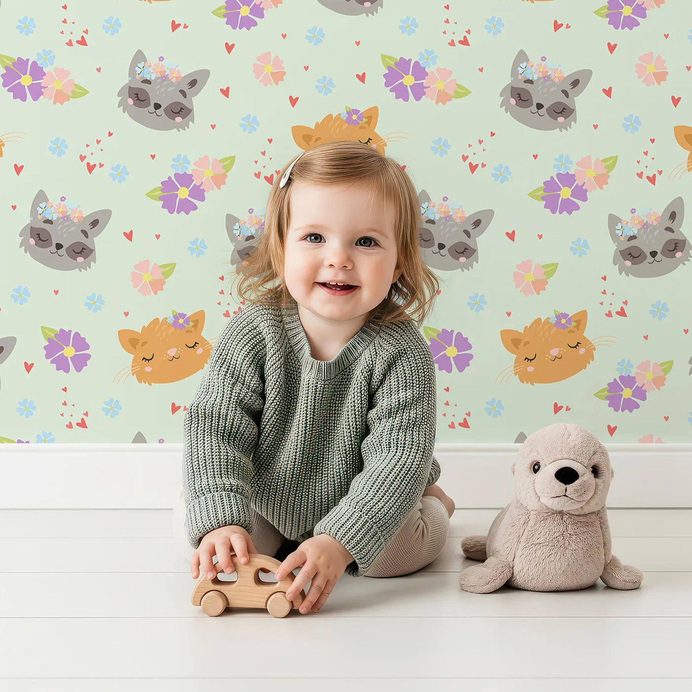 Lovely child standing in front of Whimsy Tots Meadow Snuggle Faces Wallpaper in Mint Green with cat, raccoon, flower, heart in design.
