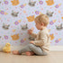 Lovely child standing in front of Whimsy Tots Meadow Snuggle Faces Wallpaper in Lilac Lavender with cat, raccoon, flower, heart in design. Lovely child standing in front of Whimsy Tots Meadow Snuggle Faces Wallpaper in Lilac Lavender with cat, raccoon, flower, heart in design.