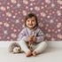 Lovely child standing in front of Whimsy Tots Twilight Wildflower Waltz Wallpaper in Plum with flower, star, leaf in design. Lovely child standing in front of Whimsy Tots Twilight Wildflower Waltz Wallpaper in Plum with flower, star, leaf in design.