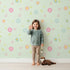 Lovely child standing in front of Whimsy Tots Breezy Blossom Meadow Wallpaper in Mint Green with flower, petal, leaf in design. Lovely child standing in front of Whimsy Tots Breezy Blossom Meadow Wallpaper in Mint Green with flower, petal, leaf in design.