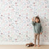 Lovely child standing in front of Whimsy Tots Fairy Petal Garden Wallpaper in White with fairy, flower, leaf, berry in design. Lovely child standing in front of Whimsy Tots Fairy Petal Garden Wallpaper in White with fairy, flower, leaf, berry in design.