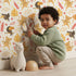 Lovely child standing in front of Whimsy Tots Monkey Banana Jungle Wallpaper in White with monkey, toucan, banana, leaf, flower in design. Lovely child standing in front of Whimsy Tots Monkey Banana Jungle Wallpaper in White with monkey, toucan, banana, leaf, flower in design.