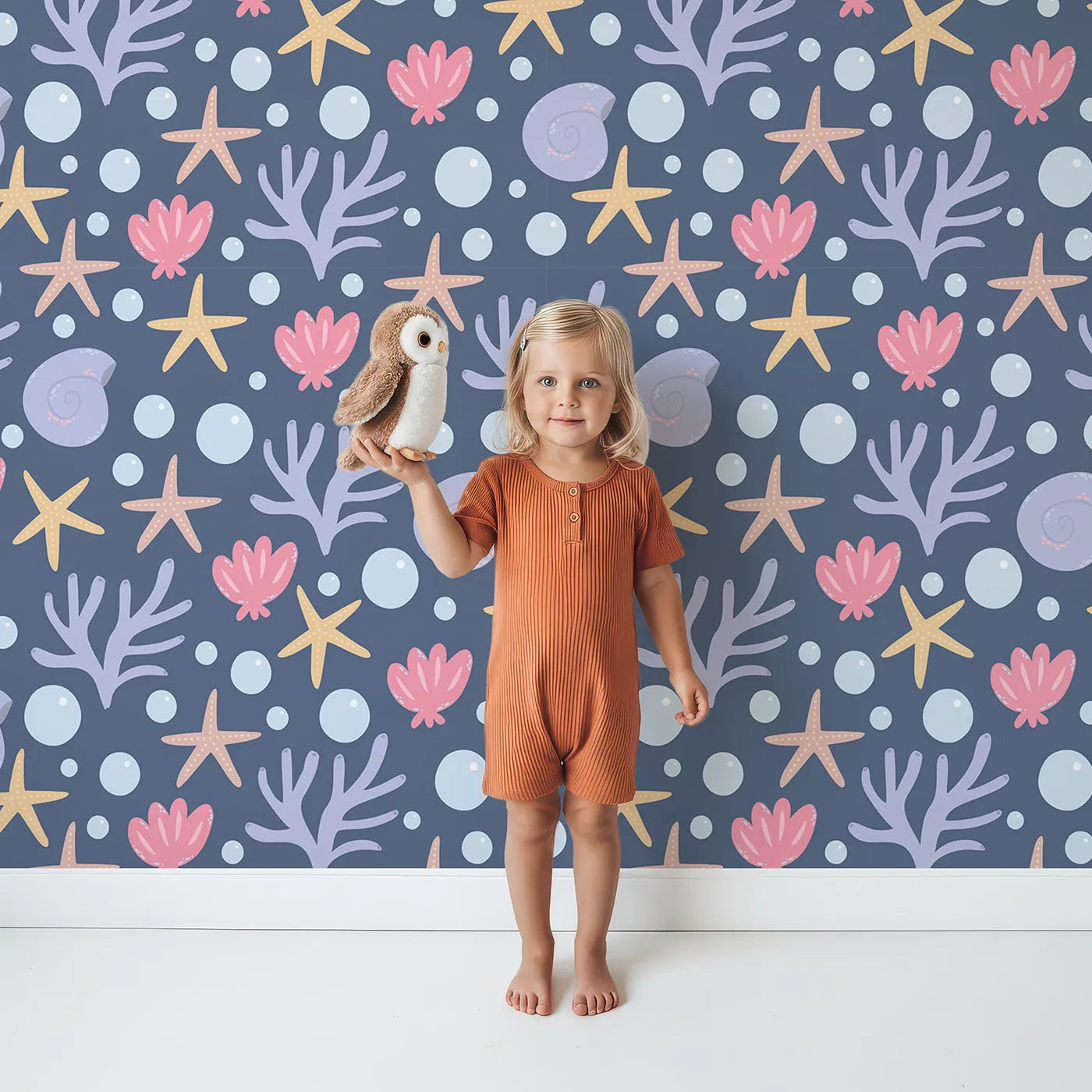 Lovely child standing in front of Whimsy Tots Starry Coral Garden Wallpaper in Deep Navy Blue with starfish, coral, shell, bubble in design.