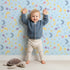 Lovely child standing in front of Whimsy Tots Starlit Wishes Wallpaper in Sky Blue with moon, star, constellation, shooting star, galaxy in design. Lovely child standing in front of Whimsy Tots Starlit Wishes Wallpaper in Sky Blue with moon, star, constellation, shooting star, galaxy in design.