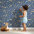 Lovely child standing in front of Whimsy Tots Starglow Constellation Dance Wallpaper in Navy Blue with moon, star, constellation, shooting star, galaxy in design. Lovely child standing in front of Whimsy Tots Starglow Constellation Dance Wallpaper in Navy Blue with moon, star, constellation, shooting star, galaxy in design.