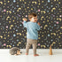 Lovely child standing in front of Whimsy Tots Starglow Constellation Dance Wallpaper in Charcoal Gray with moon, star, constellation, shooting star, galaxy in design. Lovely child standing in front of Whimsy Tots Starglow Constellation Dance Wallpaper in Charcoal Gray with moon, star, constellation, shooting star, galaxy in design.