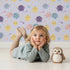 Lovely child standing in front of Whimsy Tots Planet Pals Orbit Parade Wallpaper in Lavender with planet, sun, moon, star, comet in design. Lovely child standing in front of Whimsy Tots Planet Pals Orbit Parade Wallpaper in Lavender with planet, sun, moon, star, comet in design.