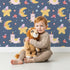 Lovely child standing in front of Whimsy Tots Llama Moon Parade Wallpaper in Soft Night Blue with llama, moon, star, flower, hat in design. Lovely child standing in front of Whimsy Tots Llama Moon Parade Wallpaper in Soft Night Blue with llama, moon, star, flower, hat in design.