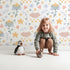 Lovely child standing in front of Whimsy Tots Starlight Blossom Dreams Wallpaper in White with star, flower, cloud, leaf in design. Lovely child standing in front of Whimsy Tots Starlight Blossom Dreams Wallpaper in White with star, flower, cloud, leaf in design.