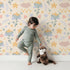 Lovely child standing in front of Whimsy Tots Starlight Blossom Dreams Wallpaper in Vanilla Cream with star, flower, cloud, leaf in design. Lovely child standing in front of Whimsy Tots Starlight Blossom Dreams Wallpaper in Vanilla Cream with star, flower, cloud, leaf in design.