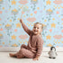 Lovely child standing in front of Whimsy Tots Starlight Blossom Dreams Wallpaper in Morning Sky with star, flower, cloud, leaf in design. Lovely child standing in front of Whimsy Tots Starlight Blossom Dreams Wallpaper in Morning Sky with star, flower, cloud, leaf in design.