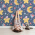 Lovely child standing in front of Whimsy Tots Starry Cuddle Sky Wallpaper in Midnight Blue with star, moon, flower, hat in design. Lovely child standing in front of Whimsy Tots Starry Cuddle Sky Wallpaper in Midnight Blue with star, moon, flower, hat in design.