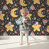 Lovely child standing in front of Whimsy Tots Starry Cuddle Sky Wallpaper in Starry Night Black with star, moon, flower, hat in design. Lovely child standing in front of Whimsy Tots Starry Cuddle Sky Wallpaper in Starry Night Black with star, moon, flower, hat in design.