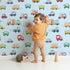 Lovely child standing in front of Whimsy Tots Bumper Buddies Rally Wallpaper in Sky Blue with car, truck, bus, foodtruck, taxi in design. Lovely child standing in front of Whimsy Tots Bumper Buddies Rally Wallpaper in Sky Blue with car, truck, bus, foodtruck, taxi in design.