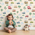 Lovely child standing in front of Whimsy Tots Bumper Buddies Forest Drive Wallpaper in Cream with car, tree, cloud, raindrop, bus in design. Lovely child standing in front of Whimsy Tots Bumper Buddies Forest Drive Wallpaper in Cream with car, tree, cloud, raindrop, bus in design.
