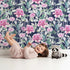 Lovely child standing in front of Whimsy Tots Rosy Garden Stroll Wallpaper in Midnight with rose, flower, stem, leaf in design. Lovely child standing in front of Whimsy Tots Rosy Garden Stroll Wallpaper in Midnight with rose, flower, stem, leaf in design.