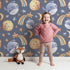 Lovely child standing in front of Whimsy Tots Starry Whale Drift Wallpaper in Indigo with whale, moon, star, comet in design. Lovely child standing in front of Whimsy Tots Starry Whale Drift Wallpaper in Indigo with whale, moon, star, comet in design.