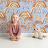 Lovely child standing in front of Whimsy Tots Rainbow Moondance Wallpaper in Blue with rainbow, moon, arc, heart in design. Lovely child standing in front of Whimsy Tots Rainbow Moondance Wallpaper in Blue with rainbow, moon, arc, heart in design.